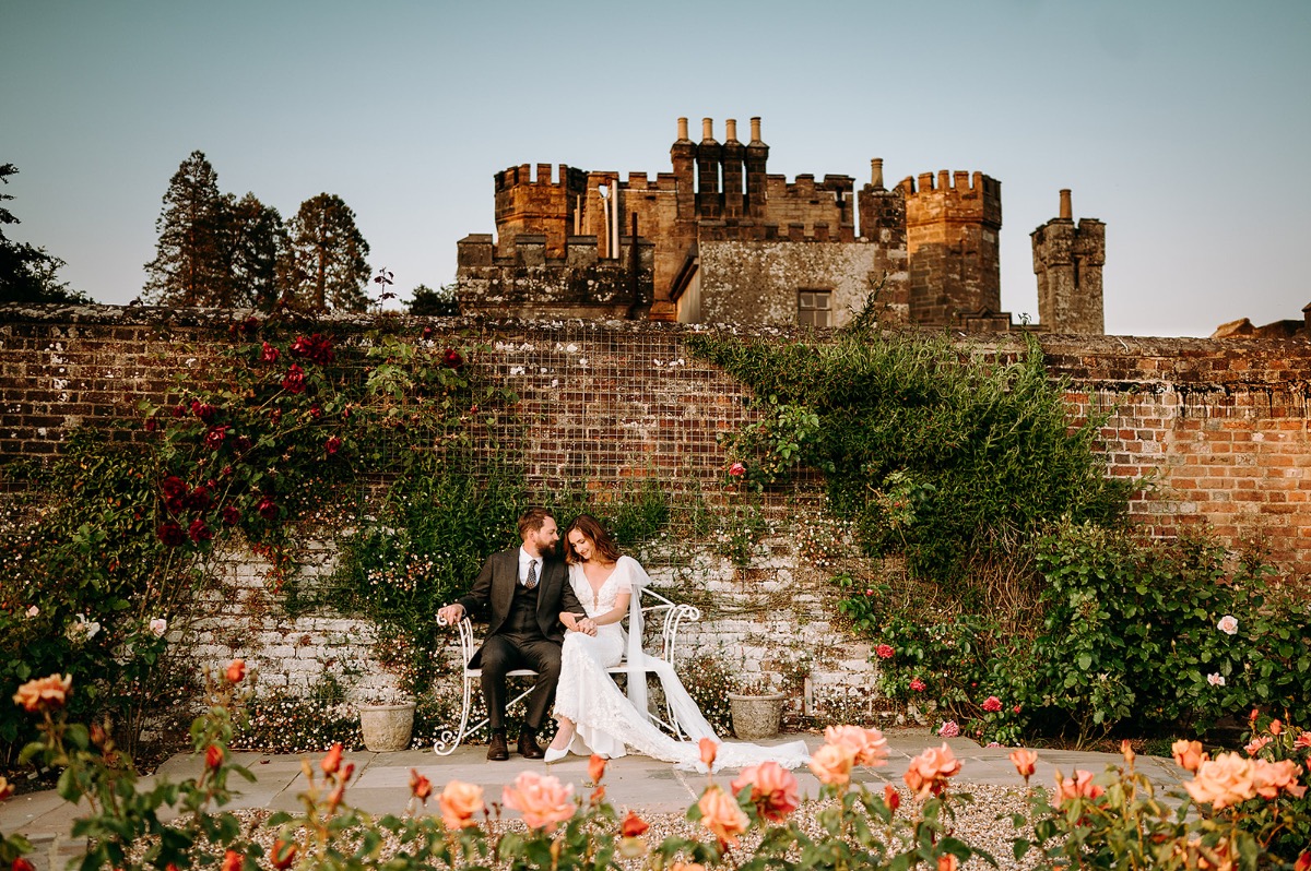 Bride and groom sitting down surrouned by flowers at golden hour with Wadhurst castle in the background