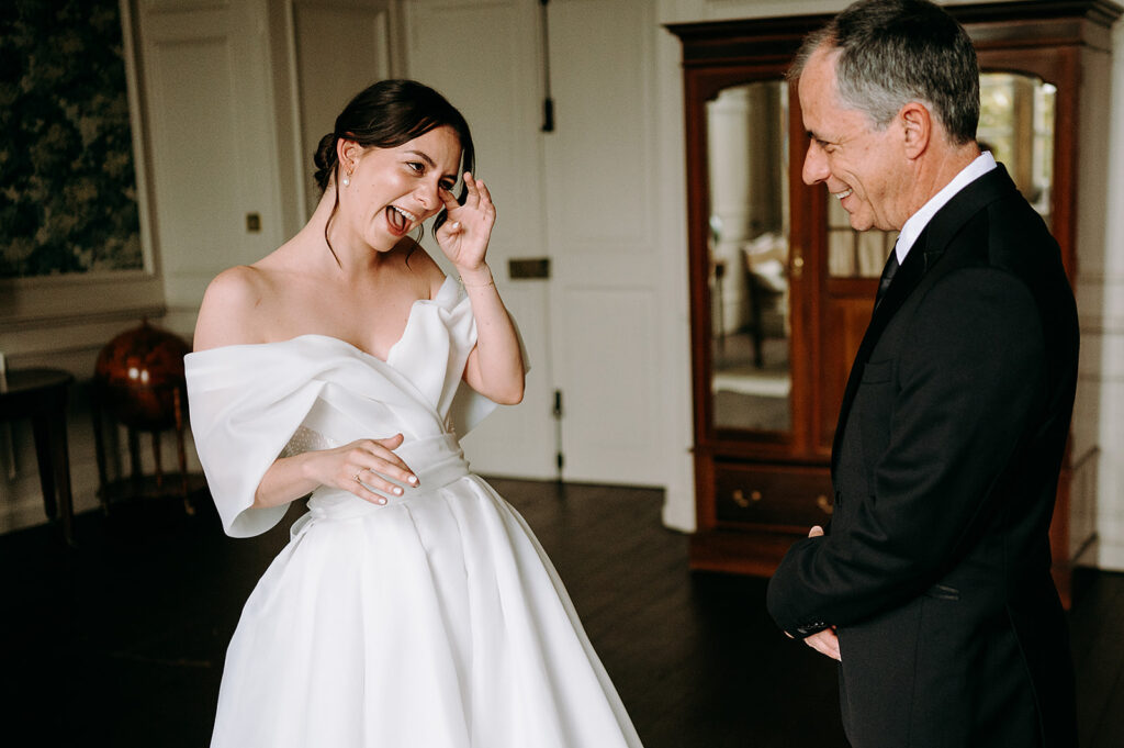 Bride in an off‑the‑shoulder white wedding dress laughs while touching her hair, facing a man in a black suit indoors with a wooden cabinet in the background.] Andrew Brannan Photography