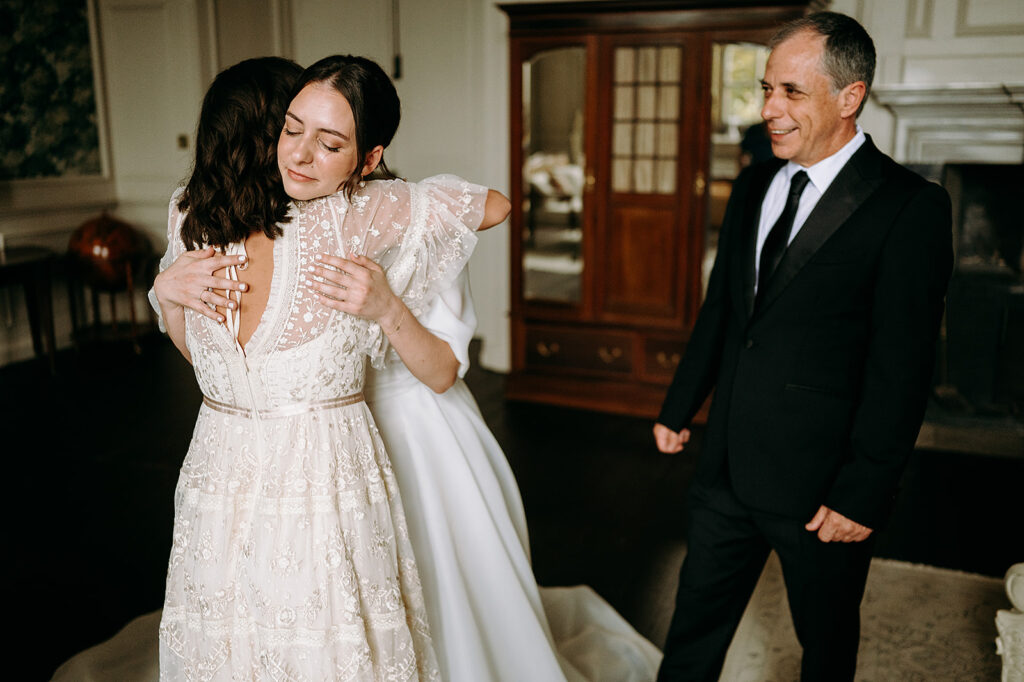 Bride in a lace wedding dress embraces a guest in a sheer-sleeved gown while a man in a black suit smiles nearby in an elegant room. Andrew Brannan Photography
