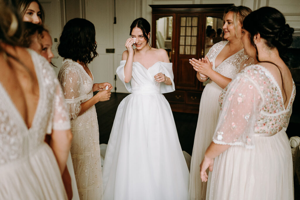 Bride in a white wedding gown wipes away a tear as bridesmaids in pale dresses applaud around her in a room. Andrew Brannan Photography