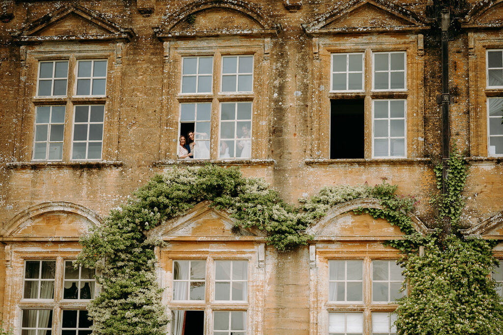 Old stone building with ivy, three women in wedding dresses leaning from a second-floor window. Andrew Brannan Photography