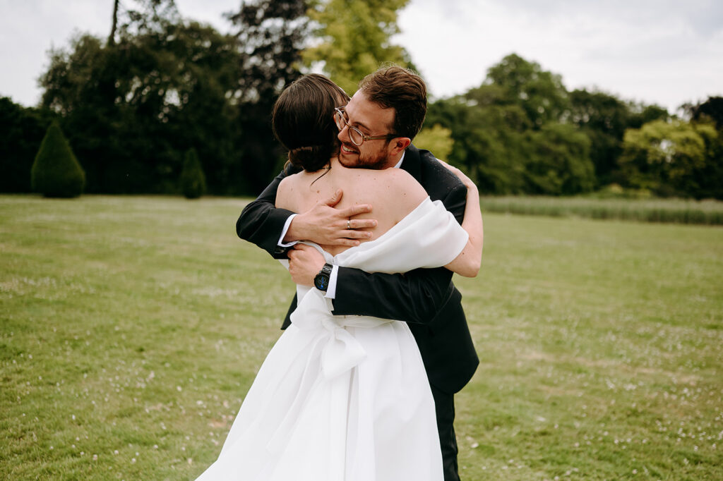 Bride in a white off-shoulder wedding dress hugs a man in a black suit in a grassy park. Andrew Brannan Photography