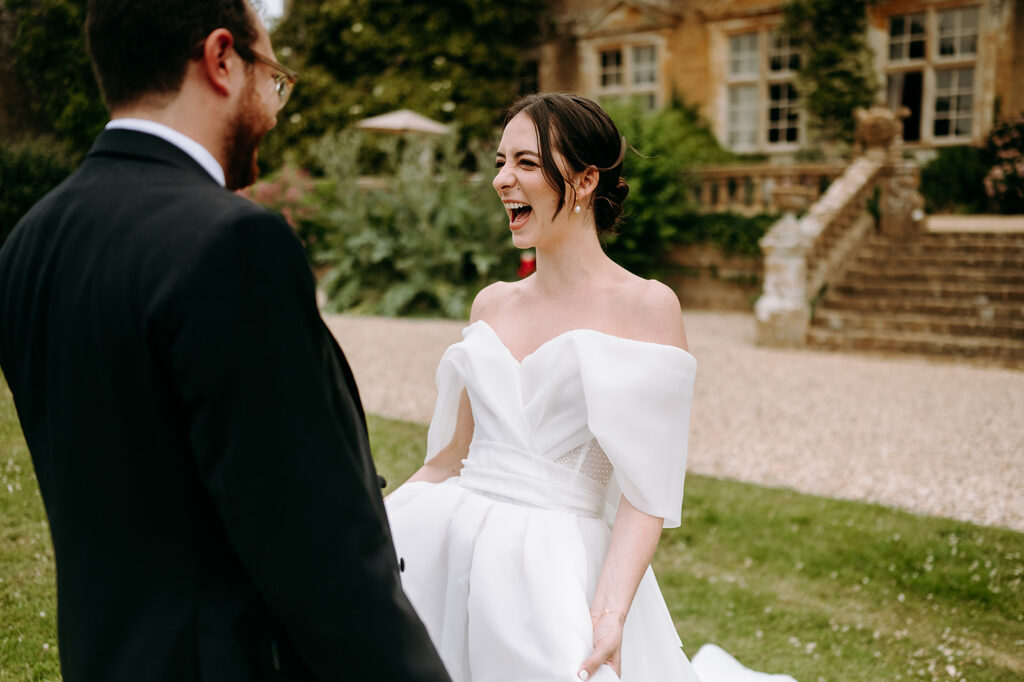 Bride in an off-shoulder white wedding dress laughs while facing a man in a black suit outdoors. Green garden and stone steps in background. Andrew Brannan Photography