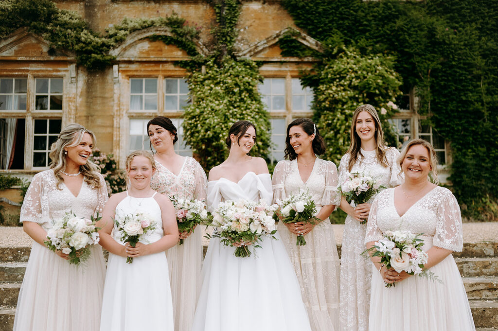 Bride in a white gown standing among six bridesmaids in pale lace dresses, each holding bouquets, on stone steps in a garden backdrop. Andrew Brannan Photography