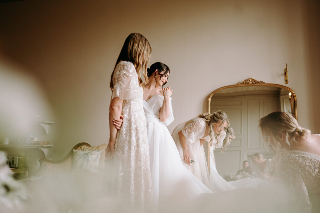 Bride in a white wedding gown standing with bridesmaids in a softly lit bridal room, preparing for the ceremony near a large ornate mirror. Andrew Brannan Photography