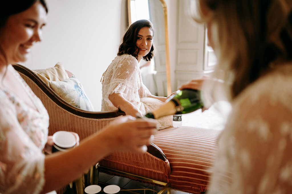 Bridesmaids in lace robes share a celebratory champagne toast in a softly lit bedroom. Andrew Brannan Photography
