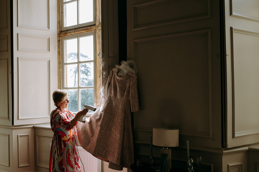 Woman in a bright patterned dress steams a long floral dress hanging by a sunlit window in a vintage, paneled room. Andrew Brannan Photography