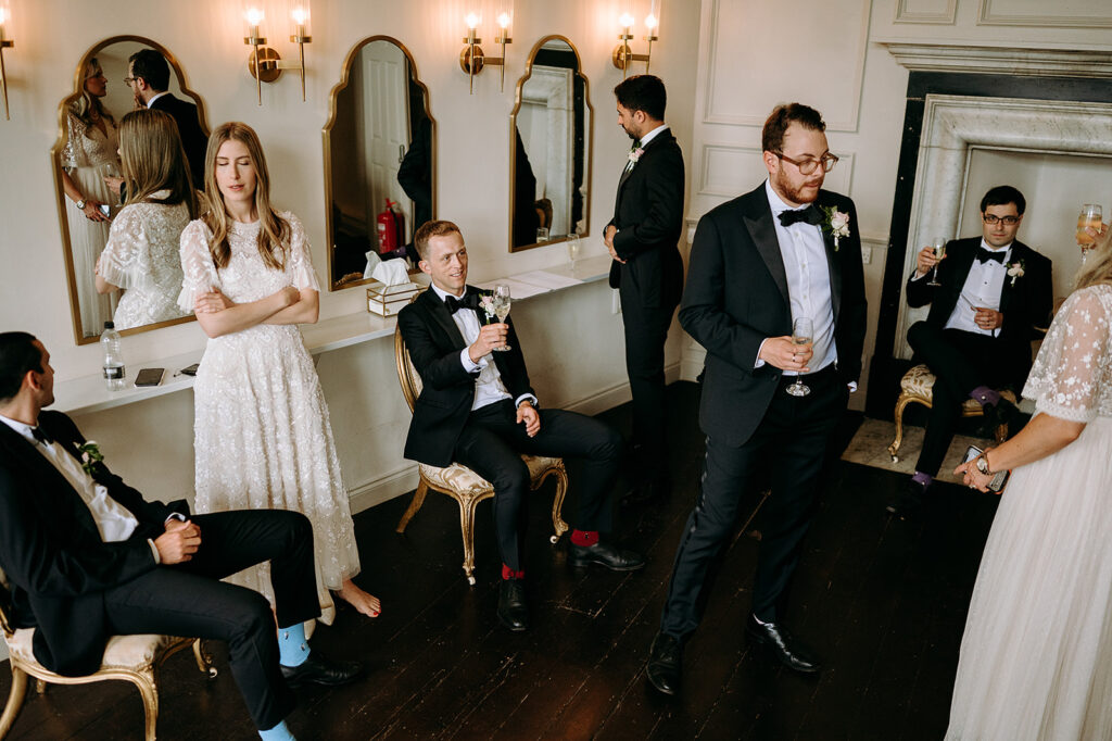 Wedding guests in formal attire mingle in a elegant dressing room, some seated and toasting with champagne Andrew Brannan Photography