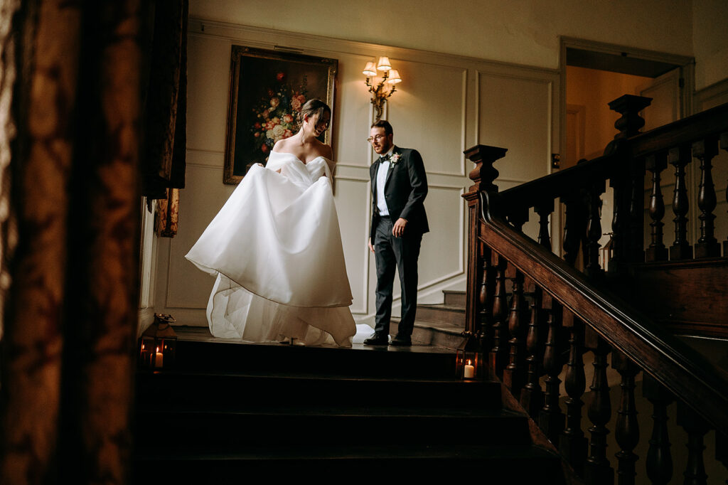 Bride lifts her voluminous white wedding gown on a grand staircase as a groom in a black tuxedo looks on in a warmly lit, elegant interior. Andrew Brannan Photography