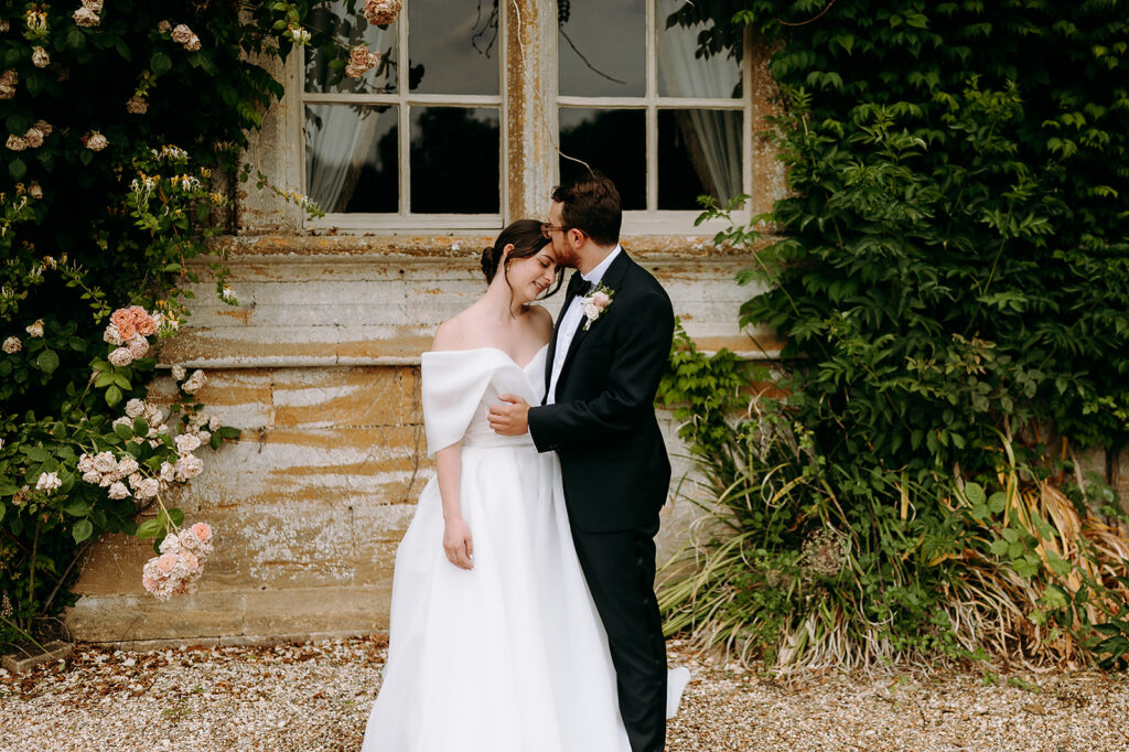 Bride in an off-shoulder white wedding gown and a groom in a black tuxedo share a tender embrace before a weathered window, surrounded by ivy and pale pink roses. Andrew Brannan Photography