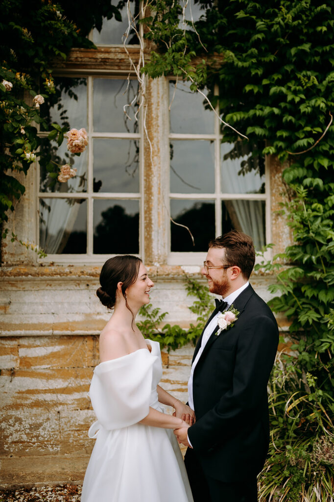 Bride in an off-shoulder white gown and groom in a black tuxedo share a smile while holding hands in front of a ivy-covered, weathered window. Andrew Brannan Photography