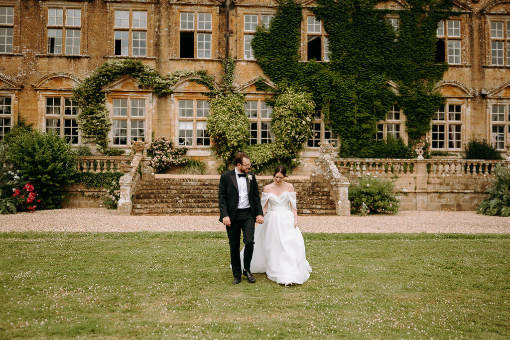 Bride in an off-shoulder white gown and groom in a black suit walk hand in hand on a grassy lawn in front of an ivy-clad historic manor. Andrew Brannan Photography