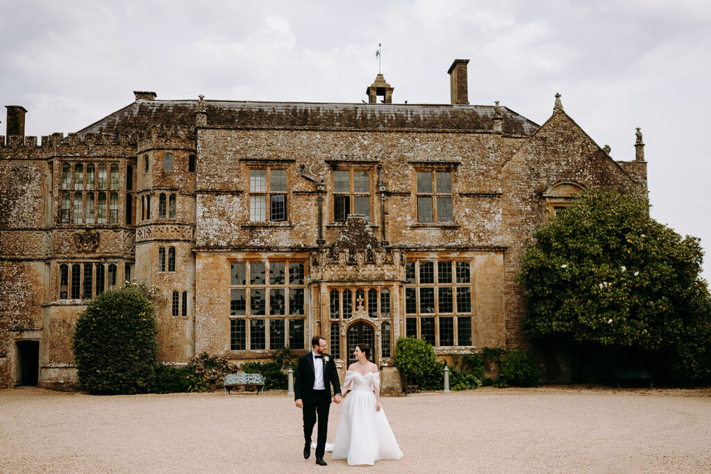A bride and groom walk hand in hand in front of a stone manor, wedding attire contrasting with the historic building. Andrew Brannan Photography