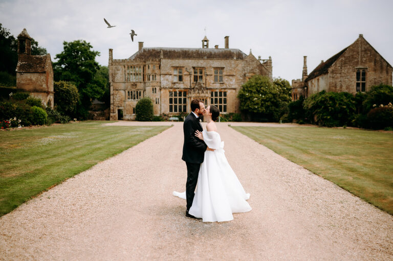 Bride in a white gown and groom in a black suit share a kiss on a gravel path in front of a historic stone manor. Andrew Brannan Photography