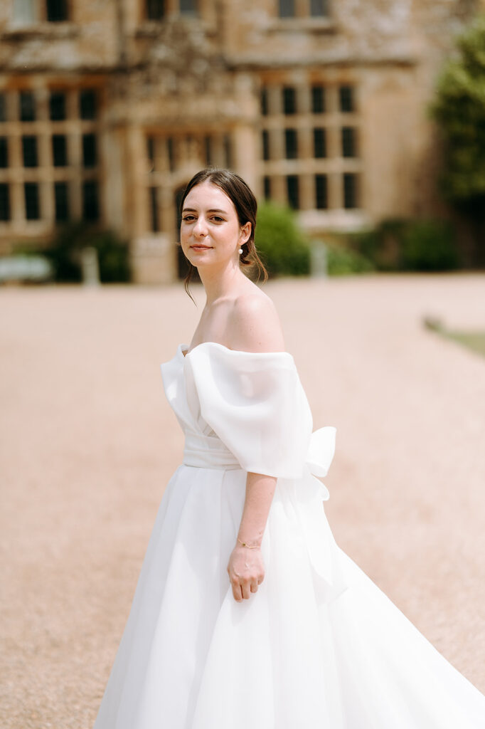 Bride in an off-the-shoulder white wedding gown standing in a gravel courtyard with a historic stone building behind her. Andrew Brannan Photography