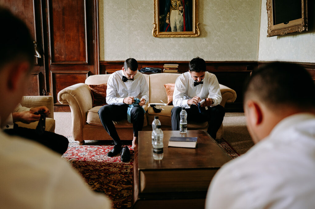 Two men in white shirts and bow ties sit on a beige sofa, concentrating on clothing items in a formal living room. Andrew Brannan Photography