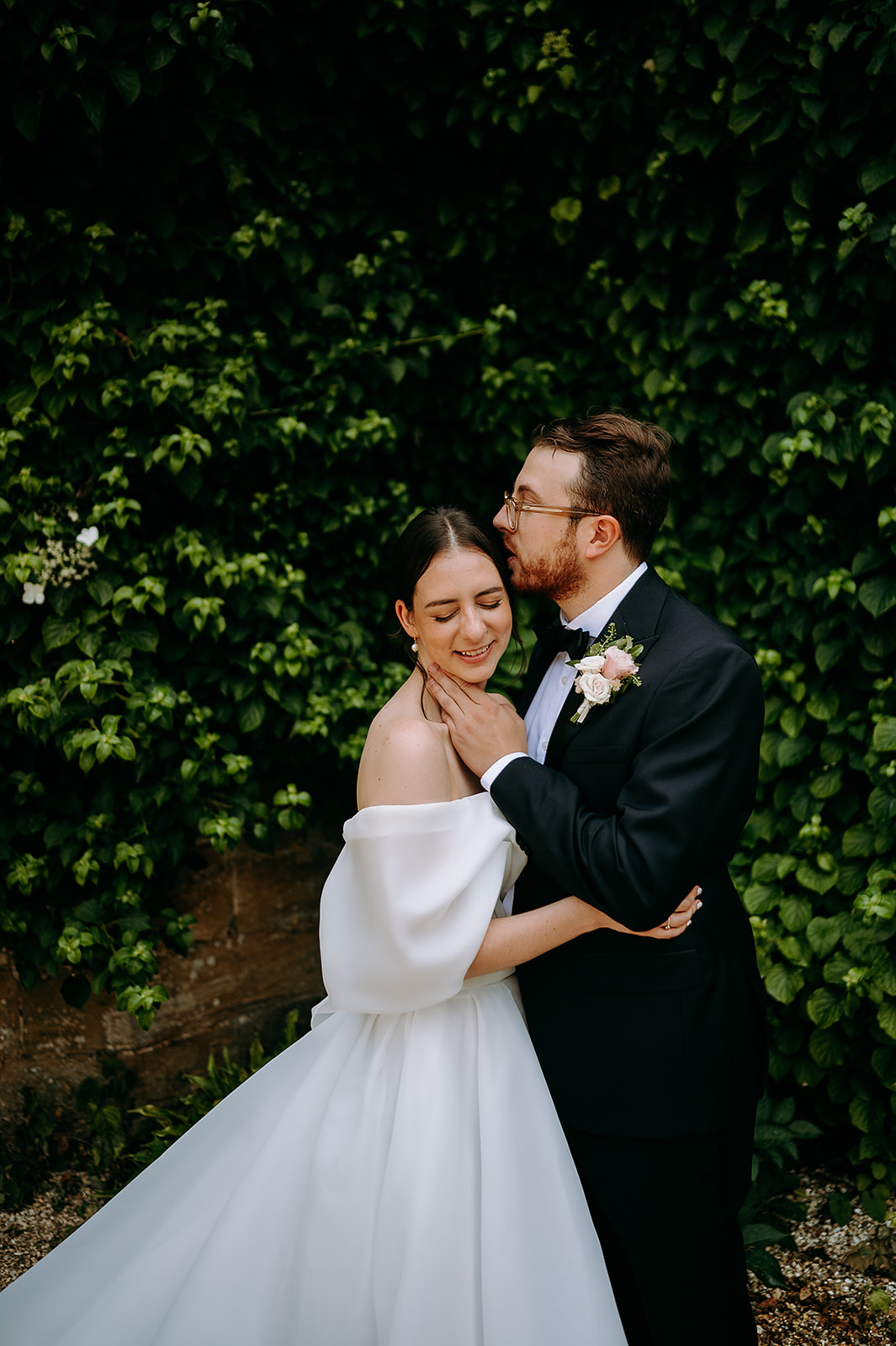 Bride in an off-shoulder white wedding dress smiles as the groom in a black tuxedo kisses her on the temple, green ivy behind them. Andrew Brannan Photography