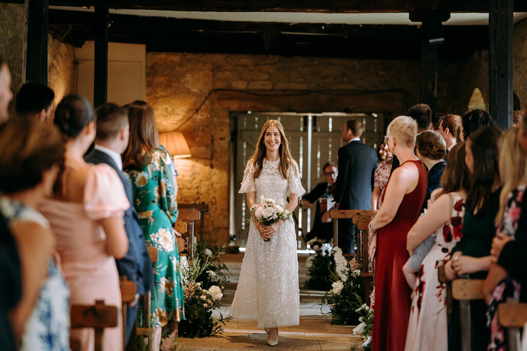 Bride in a white lace dress walks down the aisle holding a bouquet as guests look on in a rustic venue with stone walls and wooden beams. Andrew Brannan Photography