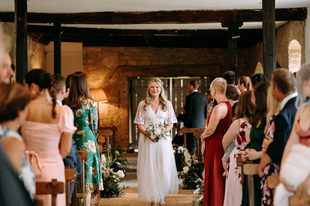 Bride in a white lace gown walks down the aisle holding a bouquet, flanked by seated guests in colorful attire. Andrew Brannan Photography