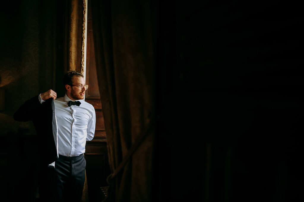 Man in a white shirt and bow tie adjusts his suit jacket by a sunlit window, wearing glasses. Andrew Brannan Photography