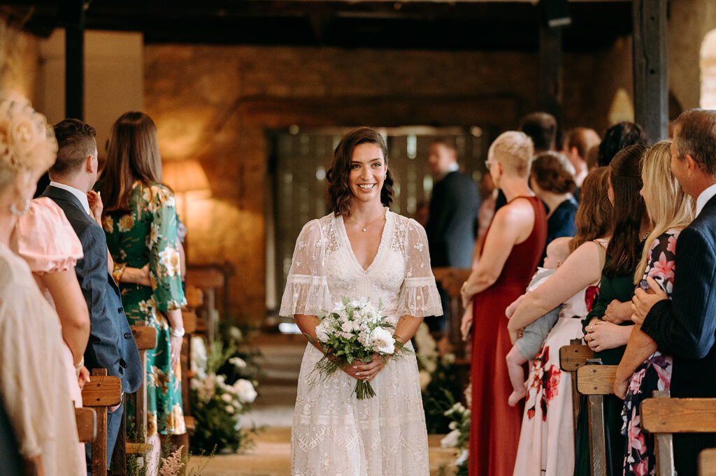 Bride in a white lace gown walking down the aisle holding a bouquet, smiling at guests on either side during a ceremony. Andrew Brannan Photography
