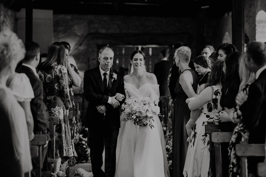 Black-and-white wedding scene: bride in an off-shoulder gown walking down the aisle escorted by a man in a suit, flanked by seated guests in formal attire. Andrew Brannan Photography