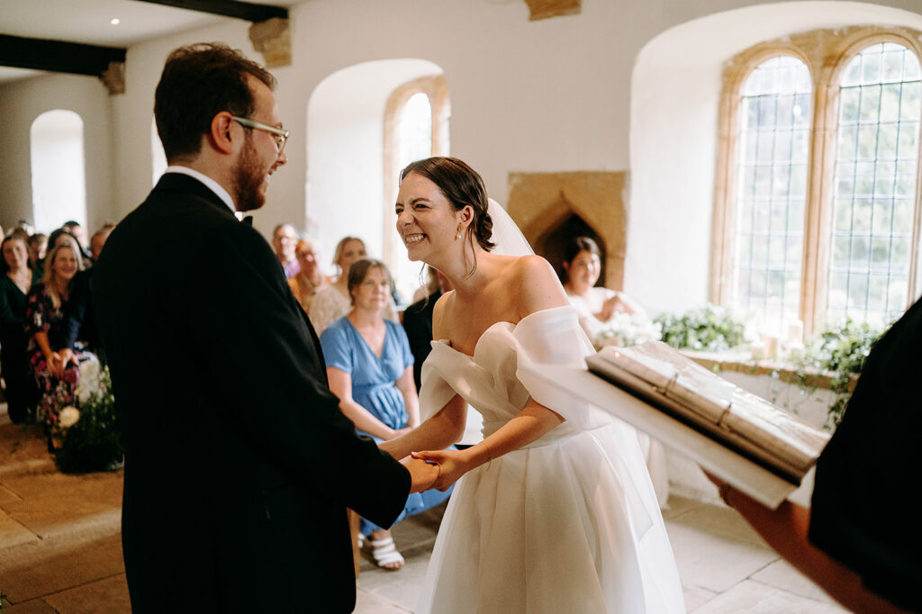 Bride in an off-the-shoulder white wedding gown smiles as she and the groom hold hands during the ceremony in a sunlit chapel. Andrew Brannan Photography