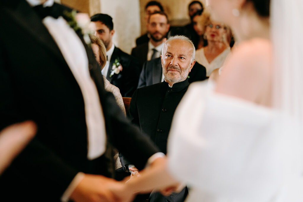 Older man in a dark suit with a gray beard watches a wedding ceremony as guests shake hands with the couple. Andrew Brannan Photography