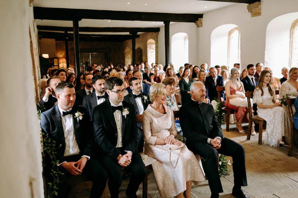 Wedding guests in formal attire seated in a sunlit historic hall with wooden beams, watching the ceremony. Andrew Brannan Photography