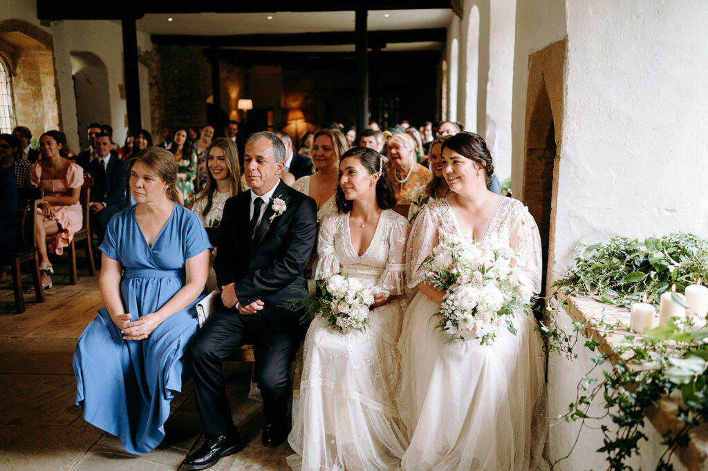 Bride in a white lace gown and two bridesmaids in light dresses sit in the front row with bouquets, during a rustic indoor wedding ceremony with guests in the background Andrew Brannan Photography