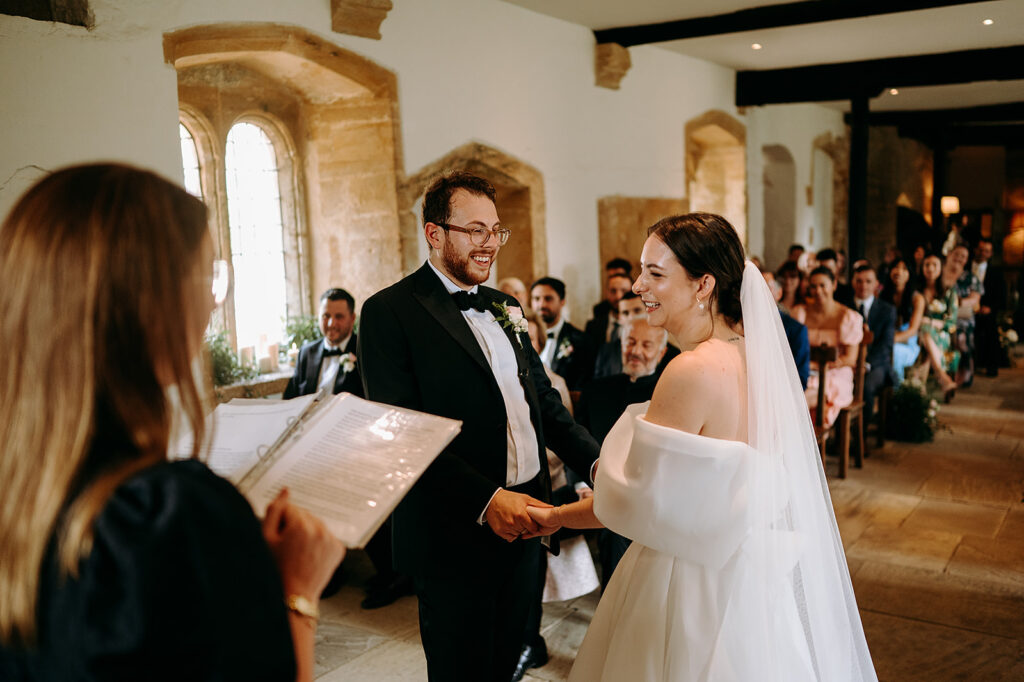 Bride and groom exchange vows at the altar, holding hands and smiling in a warmly lit chapel with guests in the background Andrew Brannan Photography