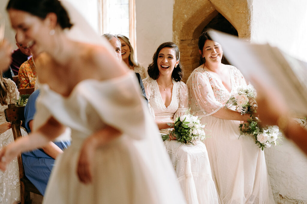 Two bridesmaids in light lace dresses sit smiling with bouquets during a wedding ceremony in a sunlit venue. Andrew Brannan Photography
