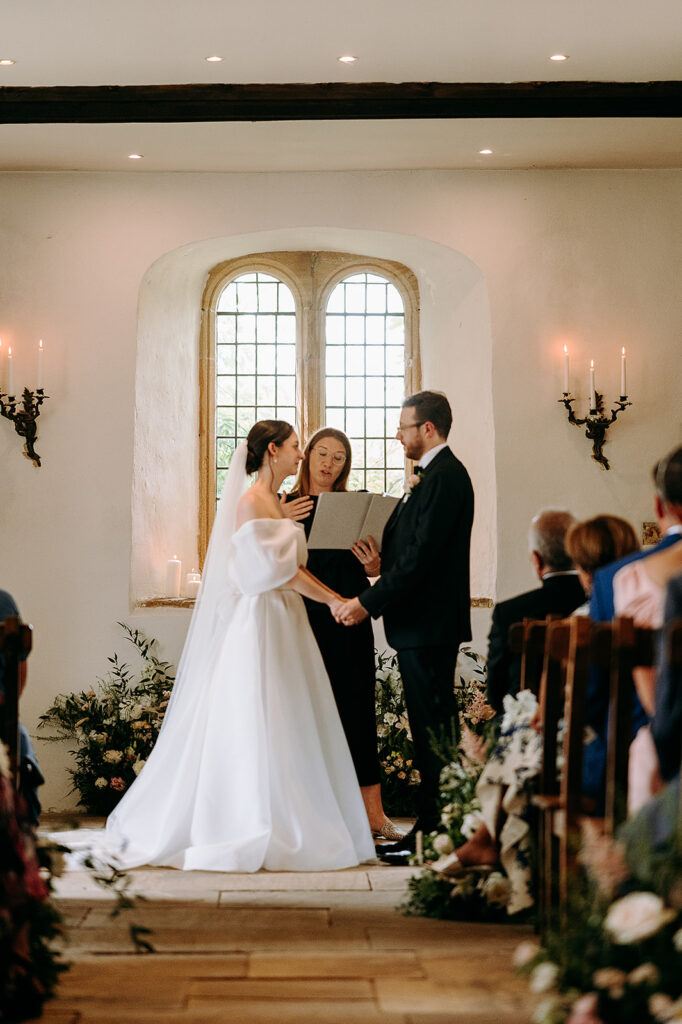 Bride and groom hold hands at the altar as the officiant reads vows in a church, with candles and flowers nearby. Andrew Brannan Photography