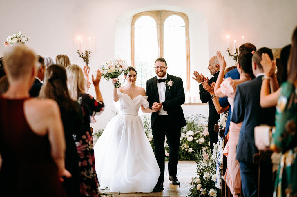 Bride in a white wedding gown holds a bouquet and walks down the aisle with her groom as guests applaud. Andrew Brannan Photography