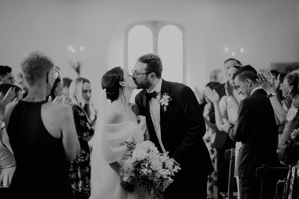Bride and groom kiss as guests applaud in a wedding aisle; bouquet in the bride’s hand, black‑tie attire throughout. Andrew Brannan Photography