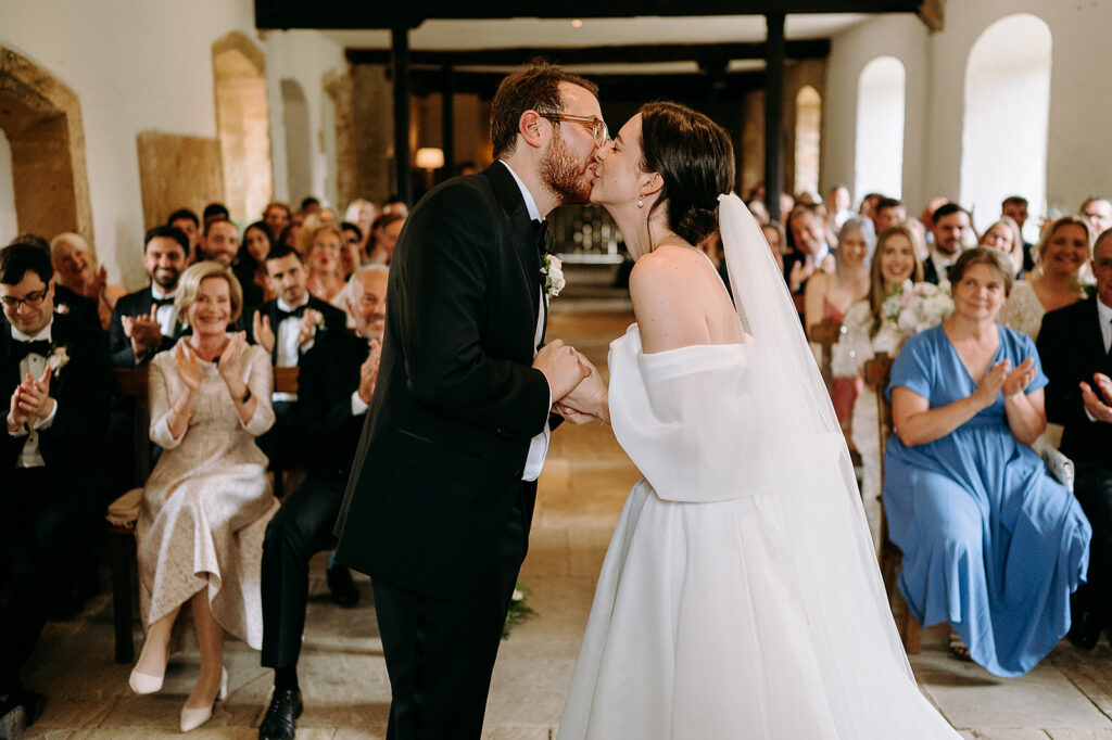 Bride and groom kiss in the aisle as guests applaud at their wedding ceremony. Andrew Brannan Photography
