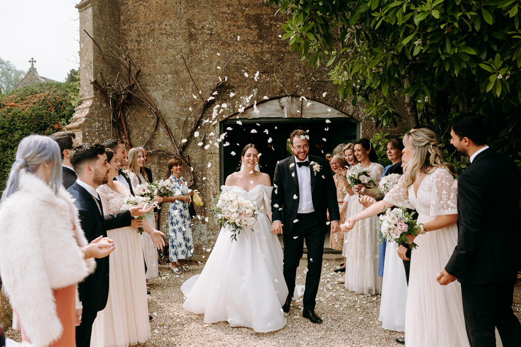 Bride and groom walk hand in hand through a line of guests throwing white petals at a rustic outdoor wedding entrance. Andrew Brannan Photography