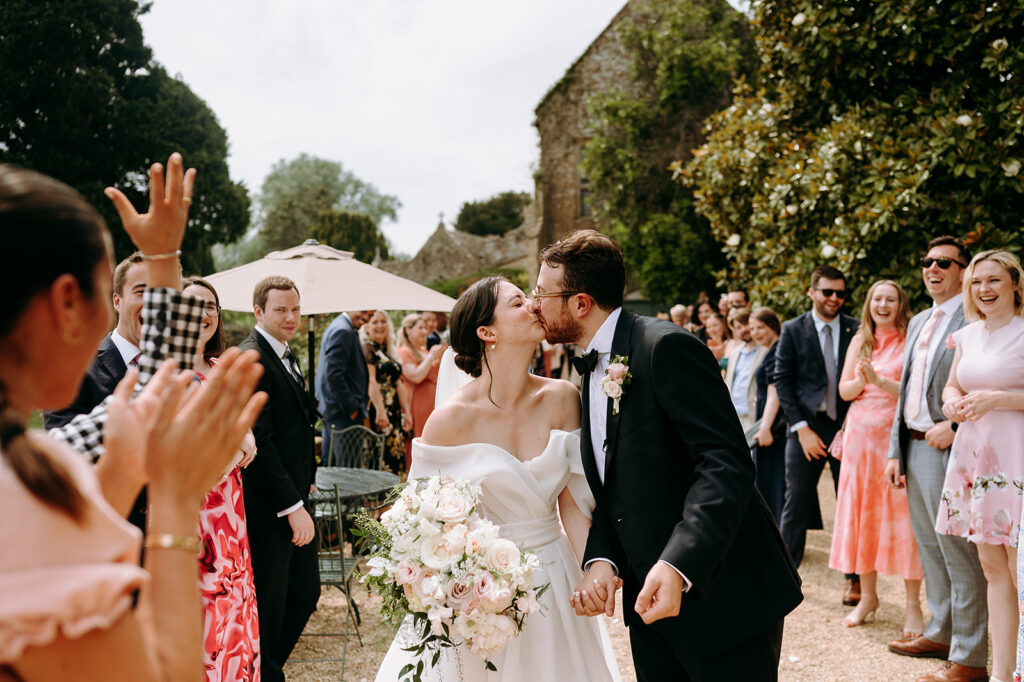 Bride and groom kiss as guests cheer at an outdoor garden wedding, holding bouquets and dressed in formal attire. Andrew Brannan Photography