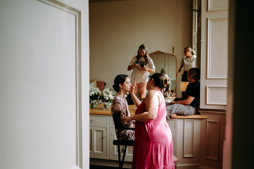 Women in a bright dressing room as one applies makeup to another, flowers arranged on a counter nearby. Andrew Brannan Photography