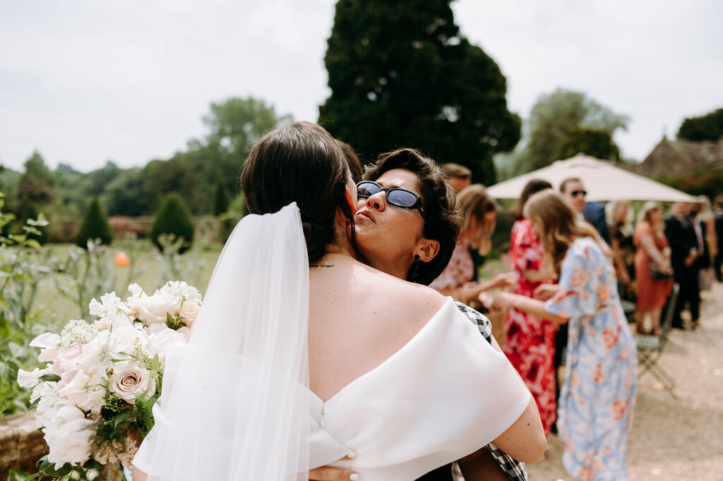 Bride in a white dress hugs a guest wearing sunglasses outdoors; pale pink bouquet in the foreground with guests mingling in the garden behind. Andrew Brannan Photography