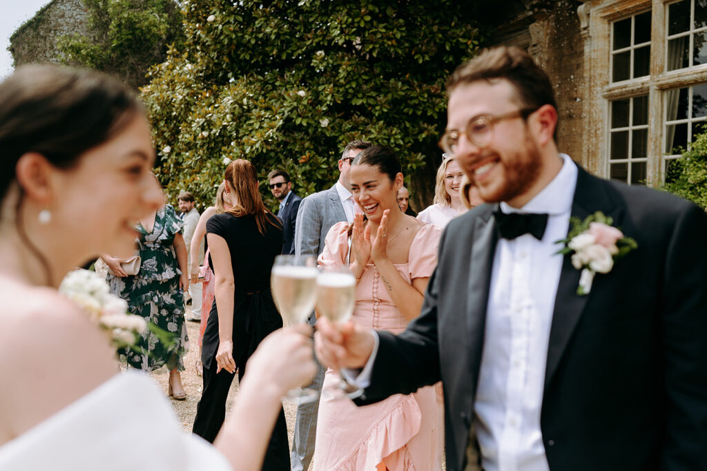Guests at an outdoor wedding toasting with champagne, smiling and chatting among greenery and a stone building backdrop. Andrew Brannan Photography