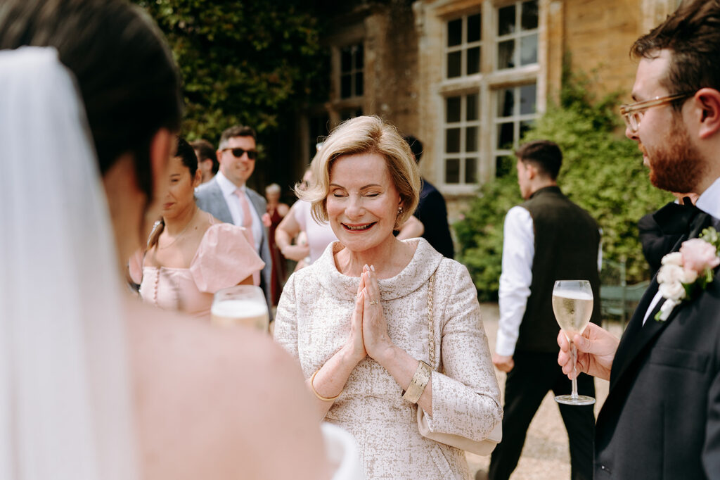 Older woman in a light patterned dress smiles with hands pressed together in a gesture of gratitude at an outdoor wedding reception. Andrew Brannan Photography