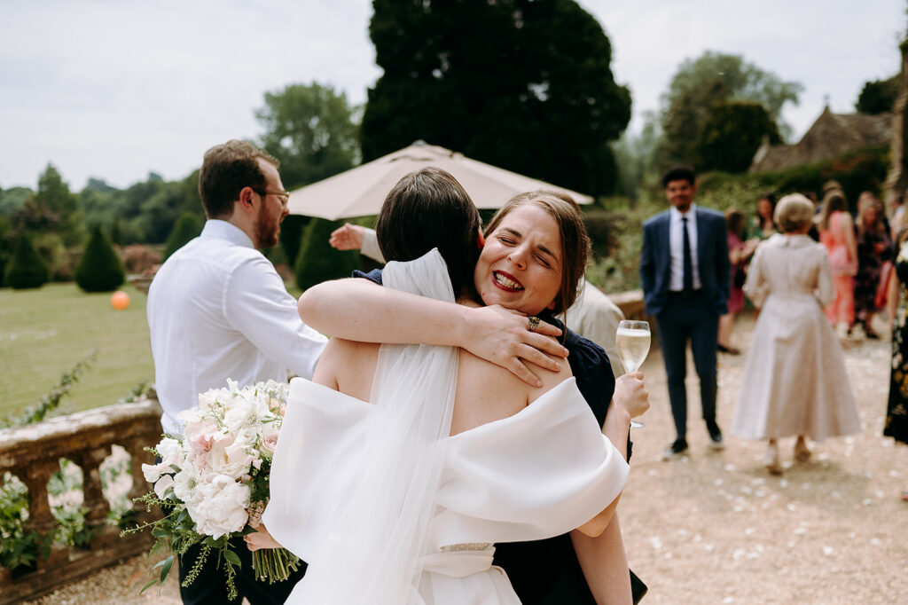 Bride in a white dress hugs a guest outdoors, bouquet in the foreground and guests mingling in the background. Andrew Brannan Photography