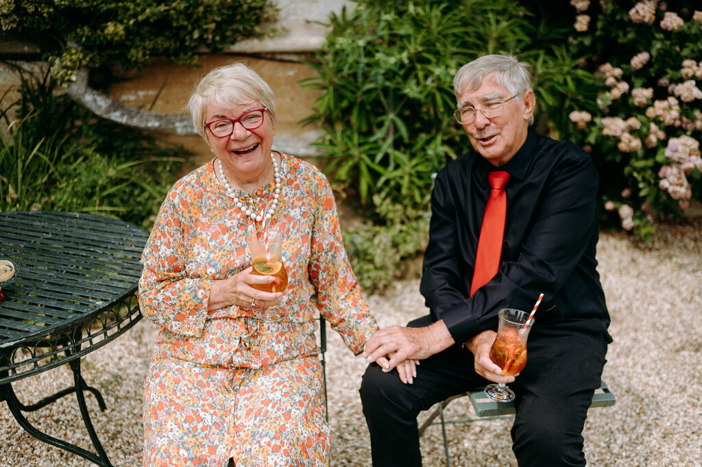 Two smiling seniors sit outdoors in a garden, each holding a glass of iced drinks; woman in a floral dress with red glasses, man in a black shirt and red tie. Andrew Brannan Photography