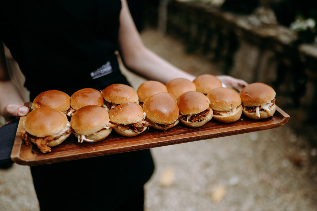 Tray of mini pulled-pork sliders on a wooden board held by a server in black attire outdoors Andrew Brannan Photography