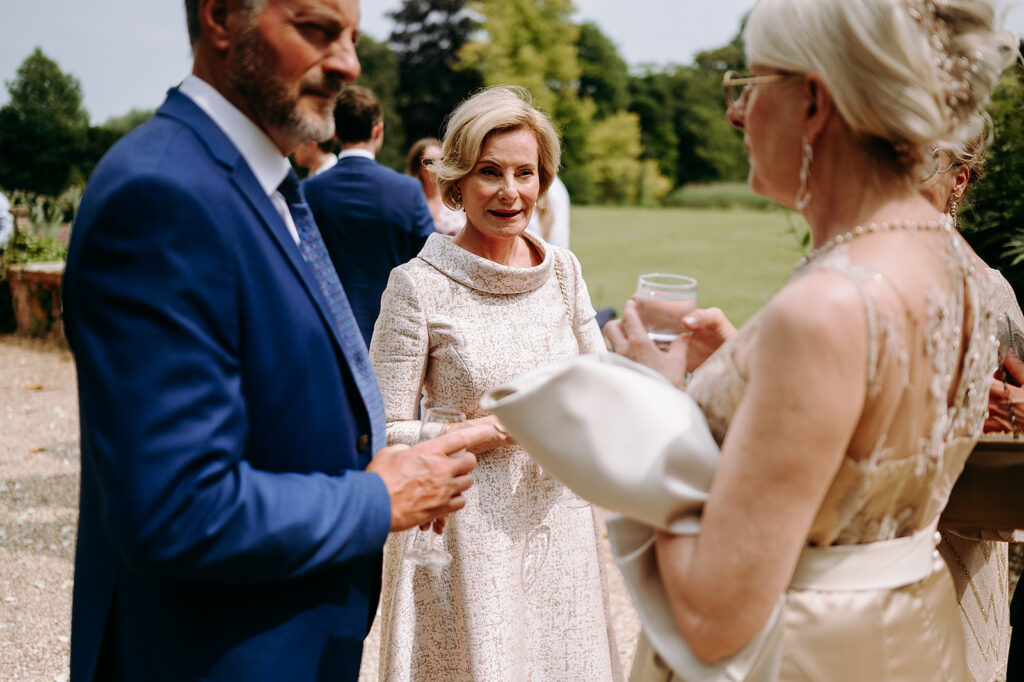 Outdoor wedding guests converse in a sunlit garden: center woman in cream lace dress, man in blue suit left, woman in champagne dress right with a glass. Andrew Brannan Photography
