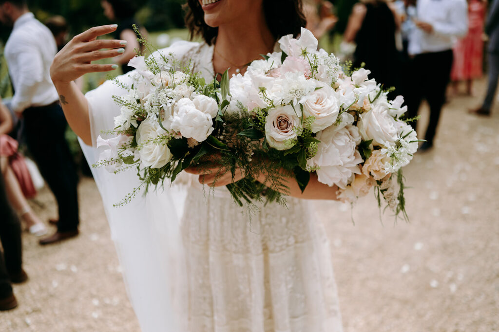 Bride holding a large bouquet of white and blush roses and peonies with greenery, in a lace dress at a wedding venue. Andrew Brannan Photography