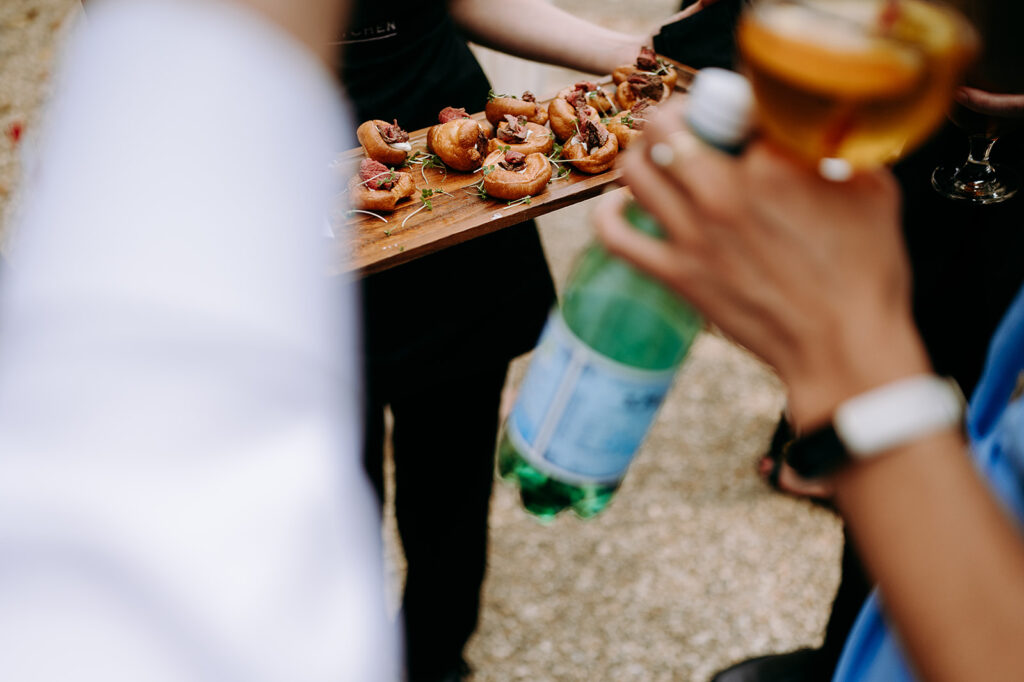 Wooden board with bite-sized meat-topped pastries being served at a party, with guests holding drinks in the foreground (blurred). Andrew Brannan Photography