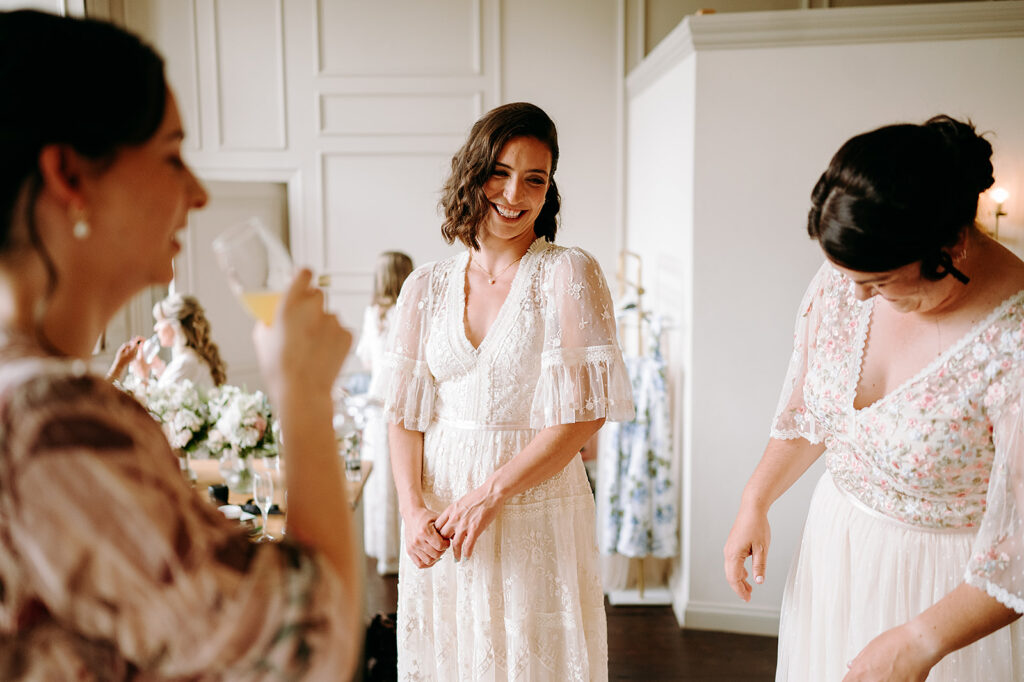Bride in a white lace wedding dress smiles as her bridesmaids help her get ready in a bright, softly lit room. Andrew Brannan Photography
