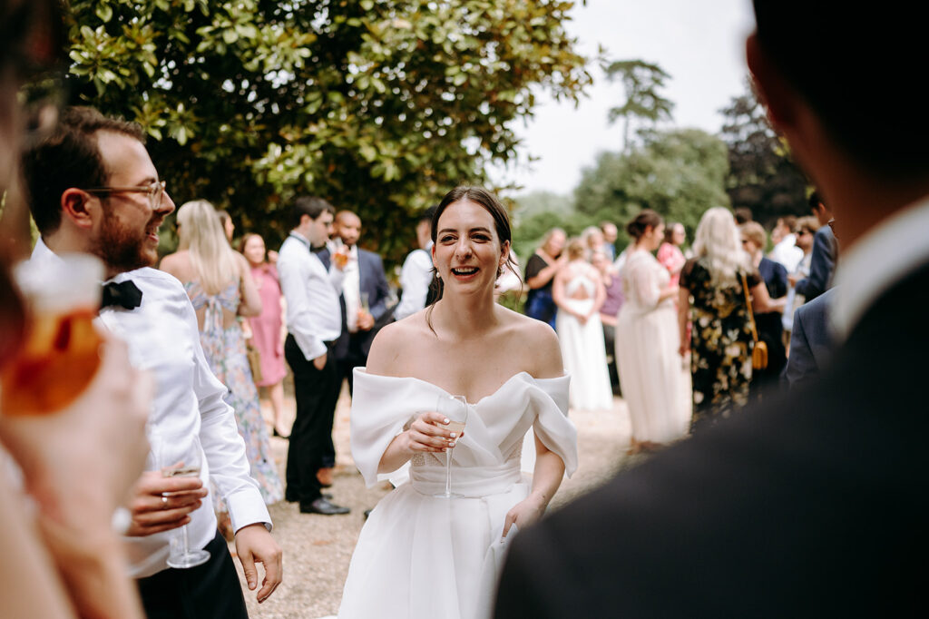 Bride in a white off‑shoulder gown smiling and holding a wine glass at an outdoor garden wedding, with guests chatting around her. Andrew Brannan Photography